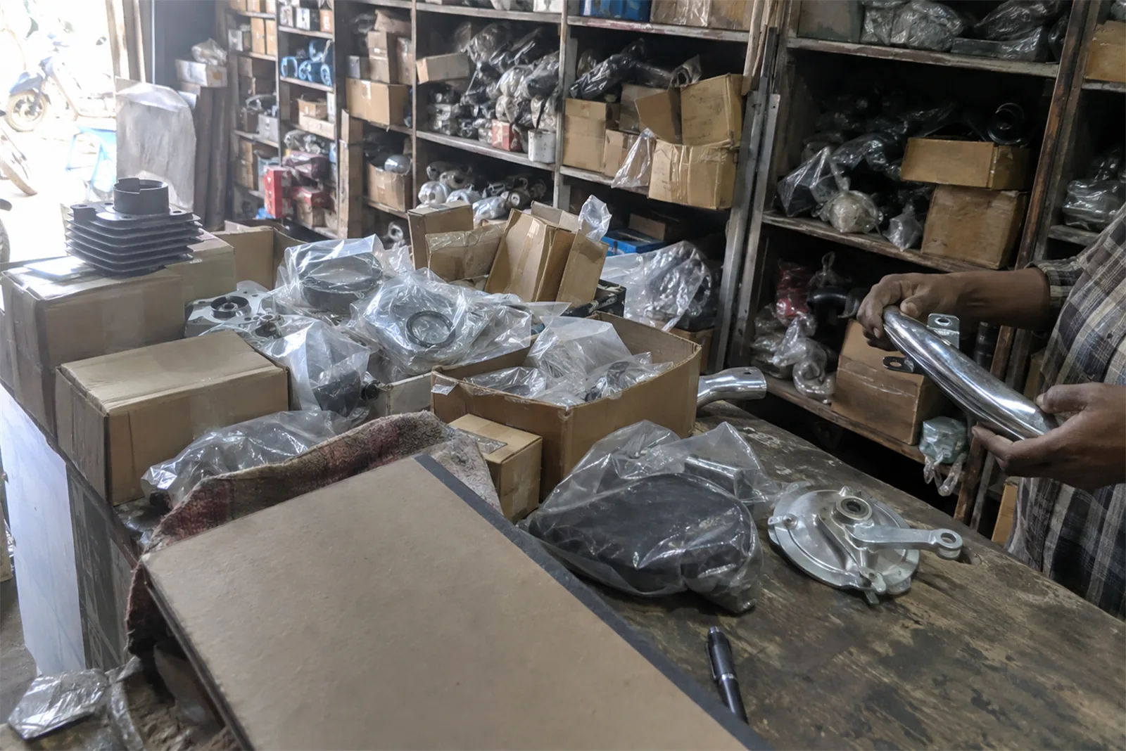 A spare parts counter stacked with two wheeler components in polythene, a chrome part held up at the shelves behind